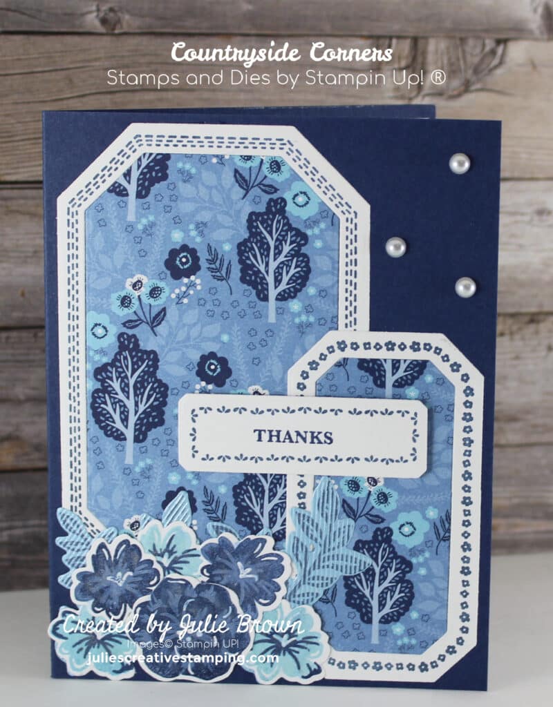 Handmade card with Night of Navy card base, Two different decorative white frames adhered to Countryside Inn Designer Series paper with trees and bushes, a decorative white sentiment label saying Thanks and various flowers and leaves stamped in Night of Navy and Balmy Blue ink cut out and layered in the bottom left corner